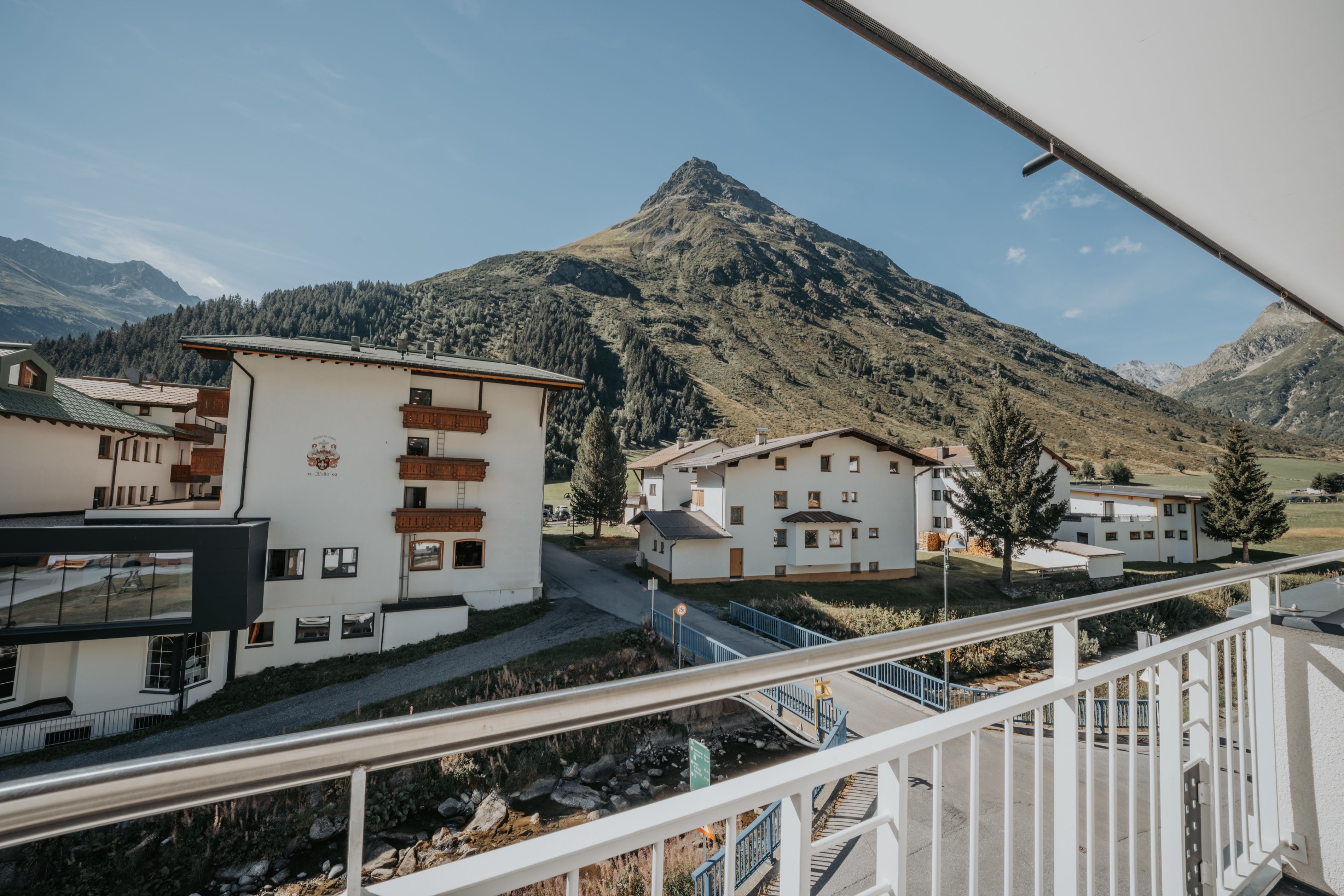Blick vom Balkon über die Dächer von Galtür auf die Ballunspitze an einem klaren Sommertag.