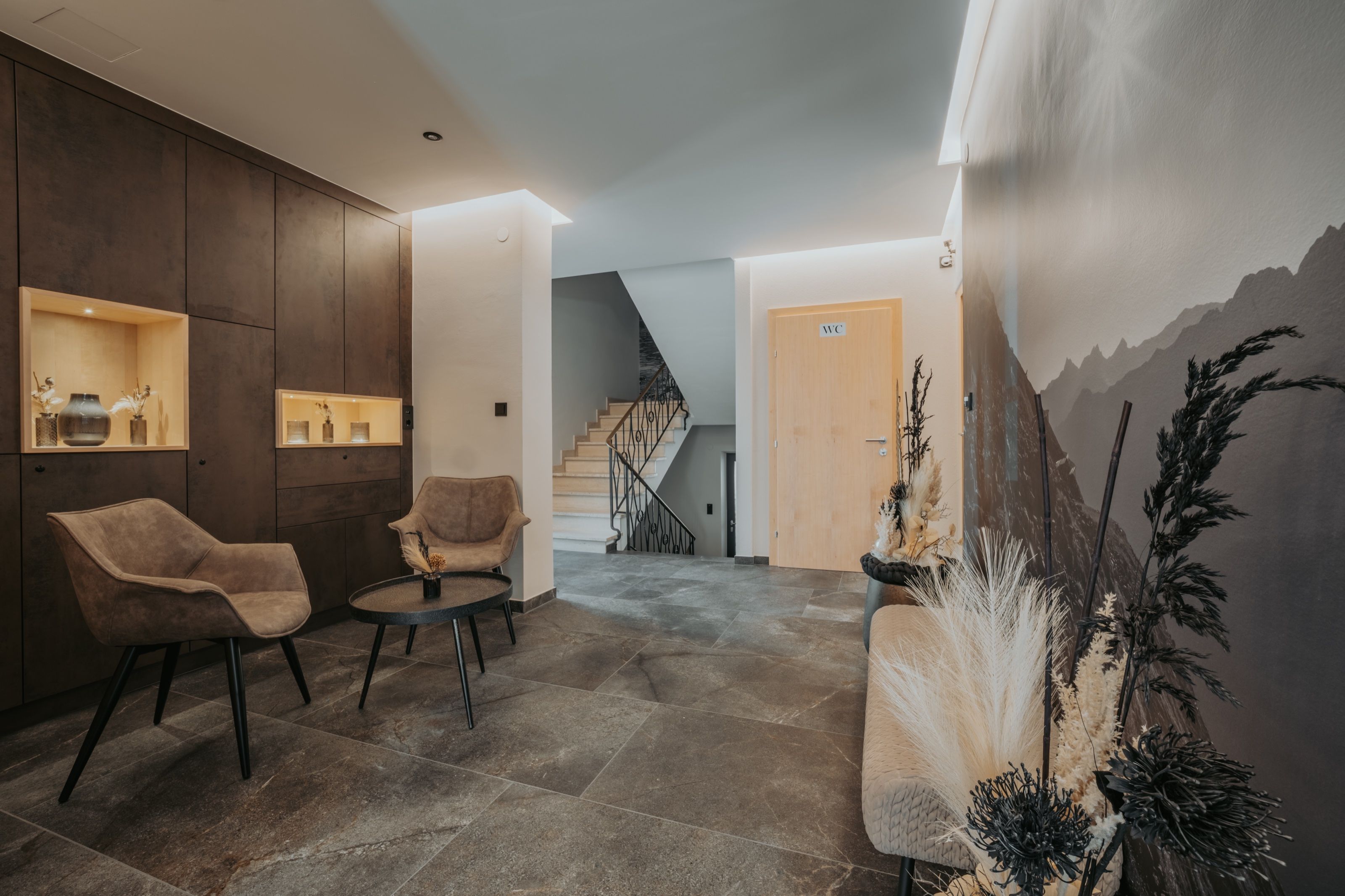 Eingangsbereich des Hauses mit zwei Polstersesseln, dunklem Steinboden und einer großformatigen Bergaufnahme an der Wand.