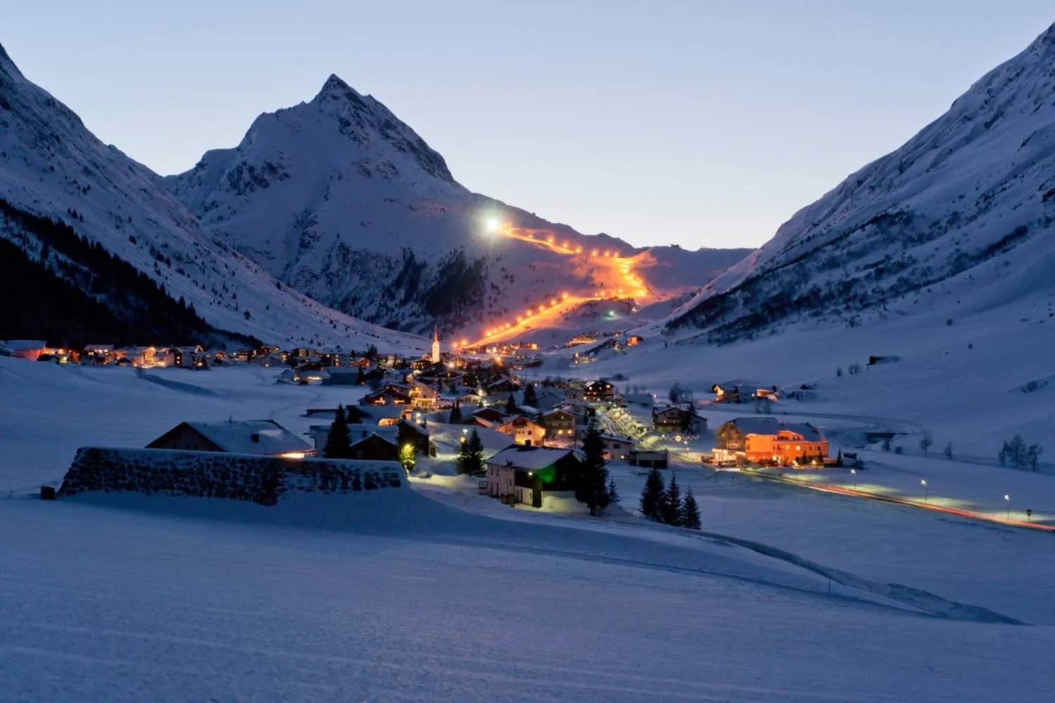 Galtür zur blauen Stunde: verschneite Häuser, warme Ortslichter und eine beleuchtete Piste am Hang.